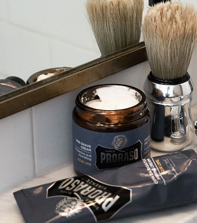 Proraso Single Blade Azur Lime Pre-Shave Cream open with Single Blade Shaving Cream Tube and Professional Shave Brush on bathroom shelf in front of mirror.