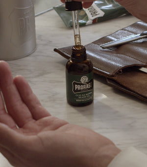 Refreshing Beard Oil  dropper being removed from bottle to be squeezed into palm of hand, on the counter of a barber shop with barber tools in the background and Refreshing Shave Cream Tube.  